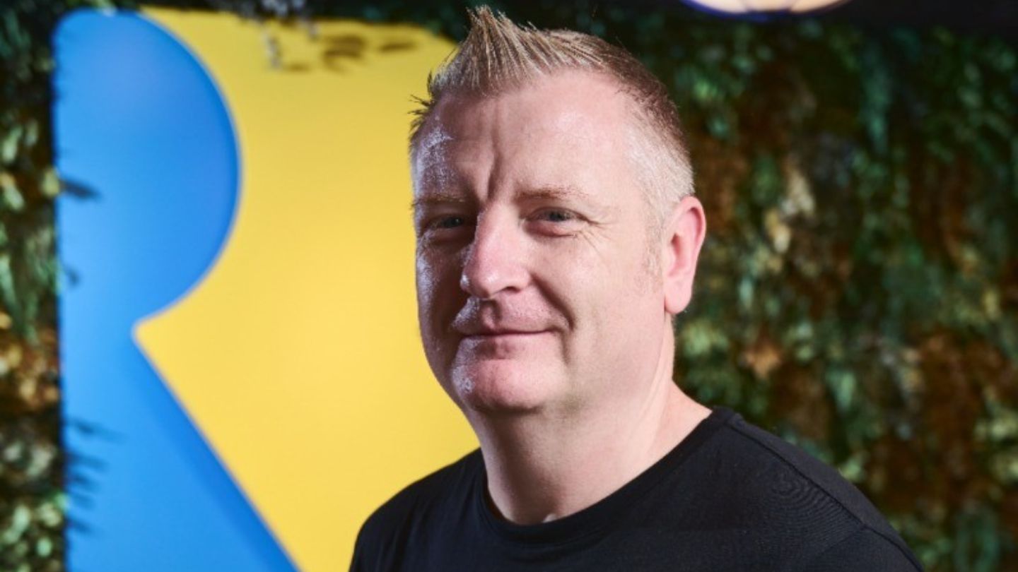 Homem sorrindo levemente posa em frente a um fundo colorido com o logotipo da Rare, estúdio conhecido por jogos ligados ao Xbox.